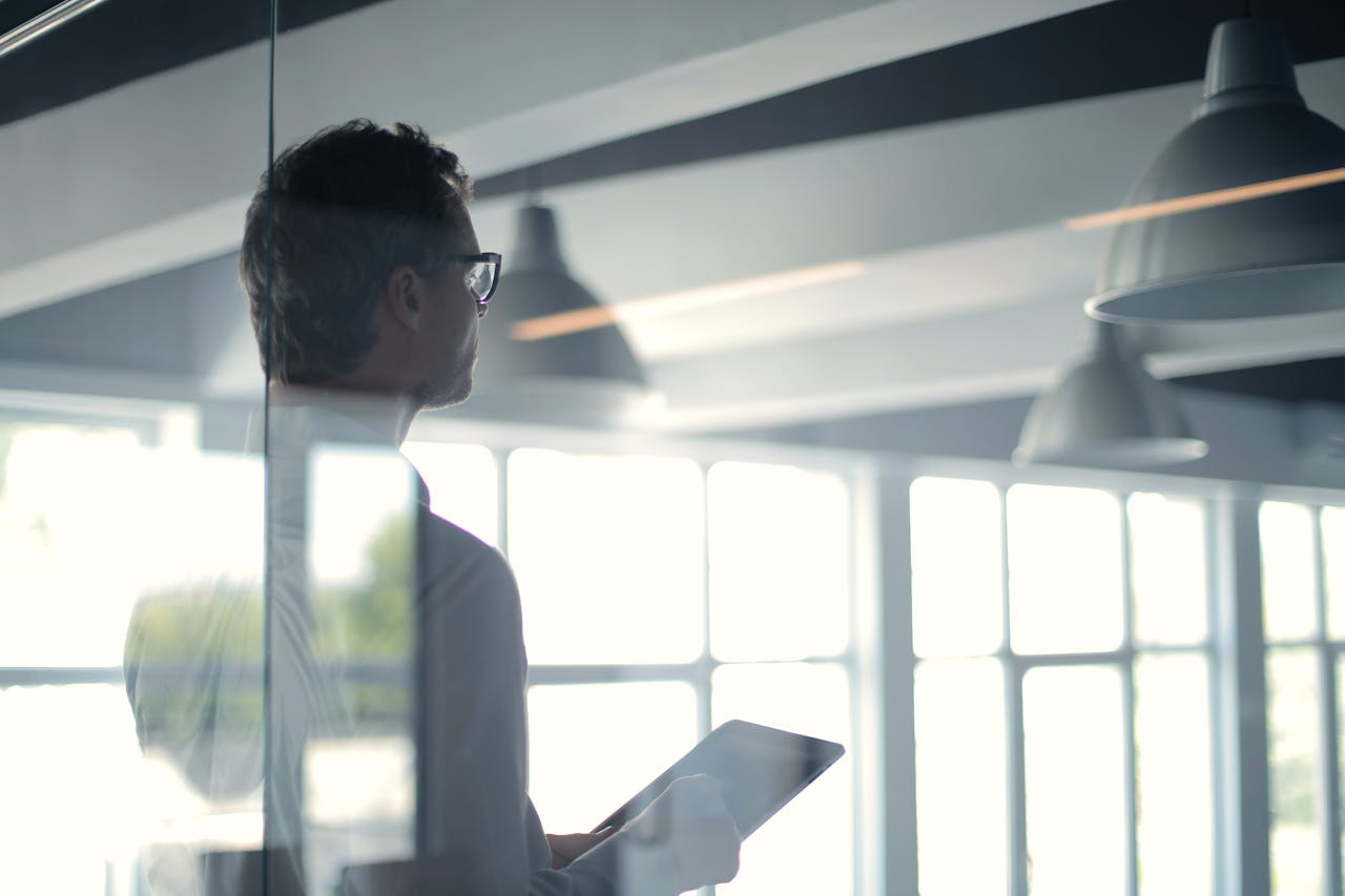 Solutions A businessman holding a tablet in an office, looking thoughtfully through a glass wall.